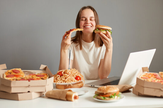Image of satisfied delighted Caucasian woman eating fast food, sitting at table with lots junk food against gray wall, holding slice of pizza and hanburger in hands, keeps eyes closed.