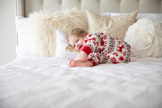 Smiling Toddler Boy Wearing Holiday Pajamas Lays on White Bed