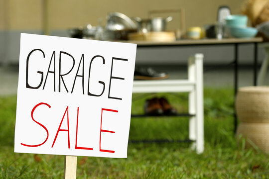 Sign Garage sale written on cardboard near tables with different stuff in yard, closeup. Space for text