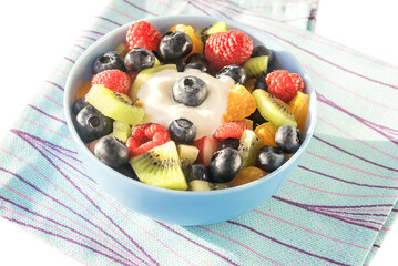 In a ceramic bowl, fresh sliced fruit salad for breakfast of raspberries, blueberries, kiwi, apples, tangerines with yogurt.