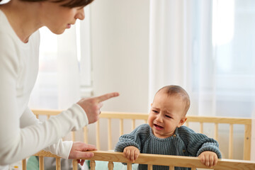 Little baby boy crying in the crib while his mother pointing finger and scolding him