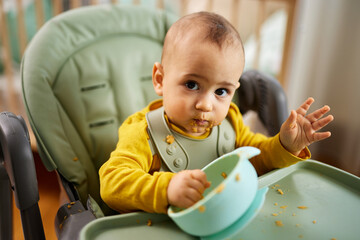 Little baby boy having his meal by himself sitting in the feeding chair at home