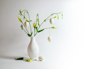 Delicate flower bunch in a white vase decorated with hanging Easter eggs on a light gray background, copy space, selected focus