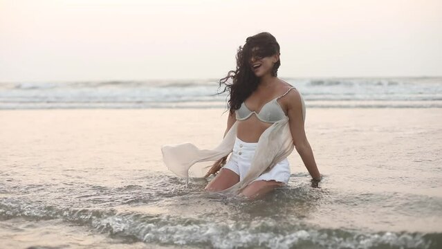 Playful beautiful woman in swimsuit and pareo sits on sand in incoming sea waves and splashes water on herself. 
