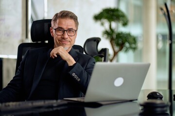 Business portrait - Businessman working with laptop computer sitting in meeting room in modern office. Happy middle aged, mid adult, mature age man smiling.