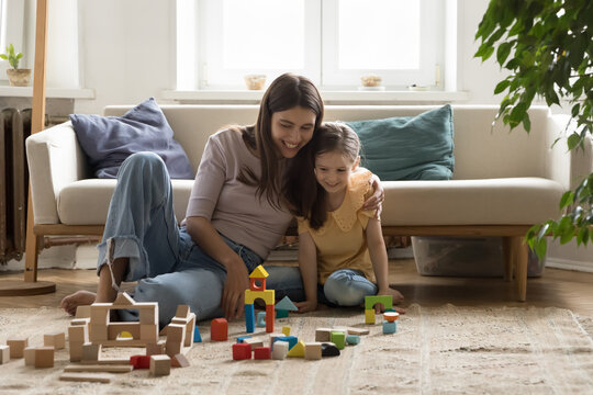 Cheerful young mom enjoying playtime with kid at home, hugging girl with proud, building toy castle from wooden constructing blocks, playing game with daughter on heating floor