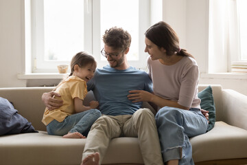 Happy young parents cuddling, tickling cheerful cute girl kid on home sofa, laughing, having fun,...