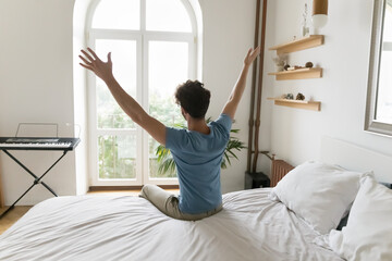 Fototapeta na wymiar Positive excited young man enjoying good morning in cozy home bedroom, sitting on bed, feeling great after sleeping enough on comfortable mattress, rising hands, looking at window. Back view