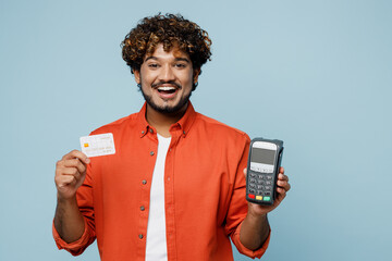 Obraz na płótnie Canvas Young fun Indian man wear orange red shirt white t-shirt hold wireless modern bank payment terminal to process acquire credit card isolated on plain pastel light blue cyan background studio portrait.