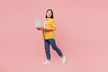 Full body side view young fun woman of Asian ethnicity wear yellow shirt white t-shirt hold use work on laptop pc computer go look aside on area isolated on plain pastel light pink background studio.