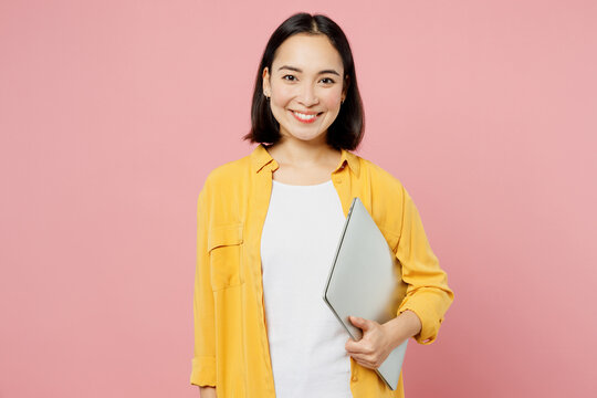 Young Fun IT Woman Of Asian Ethnicity Wear Yellow Shirt White T-shirt Hold Use Work On Laptop Pc Computer Look Camera Isolated On Plain Pastel Light Pink Background Studio Portrait. Lifestyle Concept.