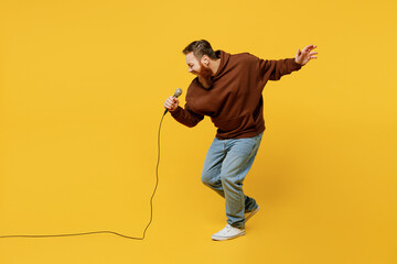 Full body happy singer fun young redhead caucasian man wearing brown hoody casual clothes sing song in microphone at karaoke club isolated on plain yellow background studio portrait Lifestyle concept - obrazy, fototapety, plakaty