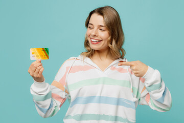 Young rich blonde woman wear striped hoody hold in hand point finger on mock up of credit bank card show thumb up isolated on plain pastel light blue cyan background studio portrait Lifestyle concept