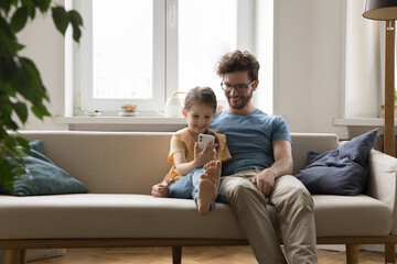 Happy dad hugging curious gen Z daughter girl using online learning app on smartphone, watching kid playing virtual game on Internet, talking on video call, resting on home sofa