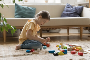 Focused cute little kid girl playing learning game on carpeted floor at home, arranging pile of colorful toy building blocks. Child constructing model from heap of colorful pieces, training creativity