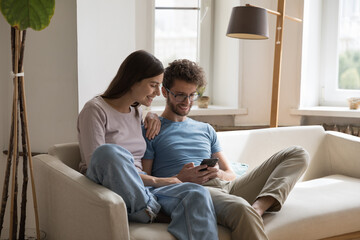 Happy young couple of husband and wife sitting on sofa together with gadget, using online app,...