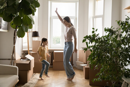 Positive active mom and little daughter kid dancing at new home, enjoying family party at stacked moving paper boxes, laughing, having fun, celebrating, house buying, relocation