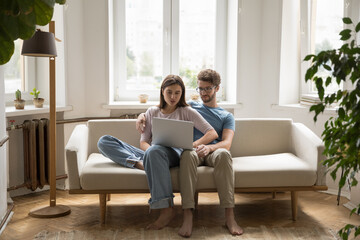 Obraz premium Young married couple using laptop at home, resting on couch with computer, shopping on Internet, booking online together, watching movie, using media service for communication
