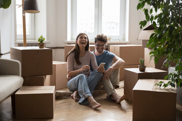Cheerful young couple of new homeowners sitting on floor at stacked moving boxes, using online app...