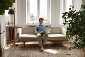 Positive freelance businessman using laptop at home, resting on couch at windows, typing, chatting,...