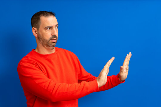 Side view of scared bearded man in beige turtleneck raising hands in sudden fear, showing stop, no, rejection gesture, looking scared, denying problem. Isolated on blue background.