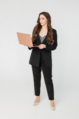 Curly adult business lady in elegant jacket standing while working on laptop against gray background