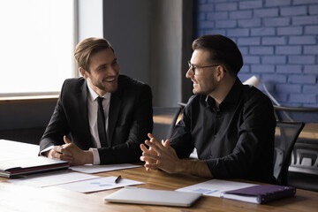 Two cheerful happy business partners men talking at work table, discussing successful startup...