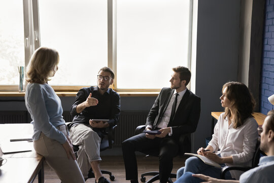 Team of employees sitting in circle in office meeting room, discussing work project, brainstorming, offering ideas, talking to senior female boss, business leader, mentor