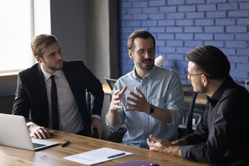 Serious confident business leader man presenting idea to colleagues, partners, speaking, telling project strategy, explaining plan, task, sitting at office work table with coworkers