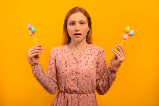 Surprised astonished shocked girl in spring floral pink dress holding multicolored painted dyed easter eggs on sticks isolated on yellow background.

Easter holiday celebration concept.