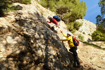two rock, cliff climbers men with backpacks climbing to top, give hand, help friend,...