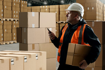 Man warehouse manager. Warehouse specialist with box. Storekeeper makes notes about arrival of goods in phone. Storekeeper in hangar. Boxes in industrial building. Man supervisor in protective helmet
