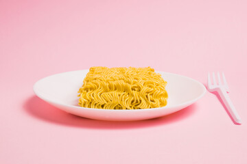 Instant noodles on a white plate with a plastic fork, on a pink background. Fast food in the modern world.