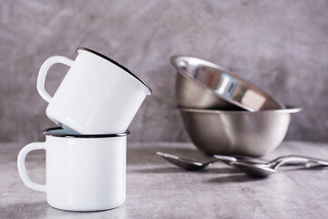 Two white empty metal mugs and metal bowls and spoons on a gray background