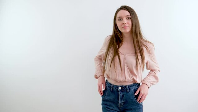 slow motion video of beautiful girl in jeans, hands in pockets on a white background, camera slowly pulls back, capturing beauty, place for text, girl moves slowly straightens hair, looks into frame