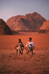 Young Couple Sitting In The Wadi Rum Desert At Sunset With Cup Of Coffee, Jordan