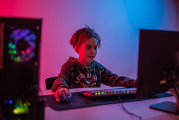 Portrait of  gamer boy playing video games on computer in dark room wearing headphones and using backlit colorful keyboard