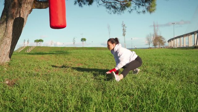 Female boxer dreesd hoodie warming up legs muscule. Athletic female prepares for boxing workout outdor in park with punching bag. Fighter warm up body before training Female fitness motivation. 