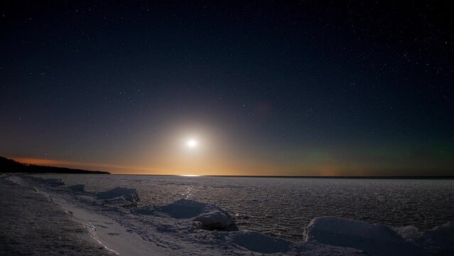 Timelapse. Seashore covered with ice under a starry sky with moon and northern lights. 