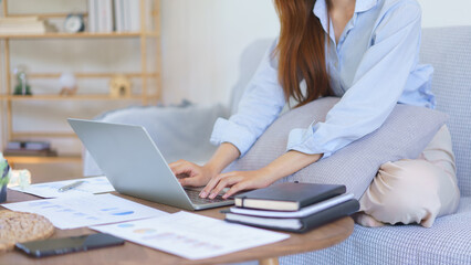 Business at home concept, Businesswoman sitting on couch to typing marketing plan report on laptop
