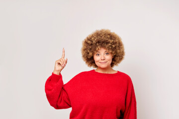 Horizontal shot of cute European young woman with curly hair points index finger up to blank space smiles cheerfully dressed in red casual pullover isolated against white background