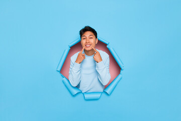 Young Asian man smiling cheerful showing and pointing with fingers teeth and mouth isolated next to blue torn wall. dental health concept. 