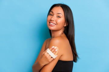 People and body care concept. Happy young Asian woman keeps body brush on the arm side body care routine smiles gladly poses over blue background