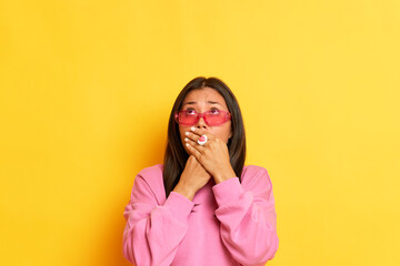 Surprised excited woman covering her mouth with both hands dressed in pink sweater wearing glasses isolated over yellow background