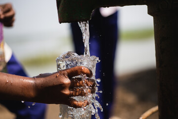 Closeup of a child hand collecting fresh drinking water from local tube-well. A hand of a kid collecting drinking water with a glass. Splash and sprinkle of water falling on glass. © Lingkon Serao