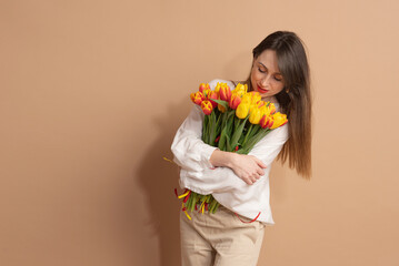 A young woman with dark hair stands on a beige background and holds a bouquet of red-yellow tulips...
