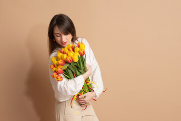 A young woman with dark hair stands on a beige background and holds a bouquet of red-yellow tulips in her hands. International Women's Day