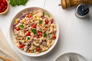 Homemade macaroni salad with elbow pasta and canned tuna, carrot, Purple cabbage, tomato, corn, green peas and mayonnaise dressing in a white plate on wooden table.Top view