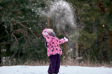 Six year old girl dressed in a blue coat and a pink hat and boots throws snow up