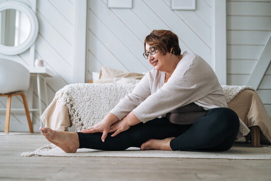 Middle aged plus size smiling brunette woman in home wear doing stretching in bedroom